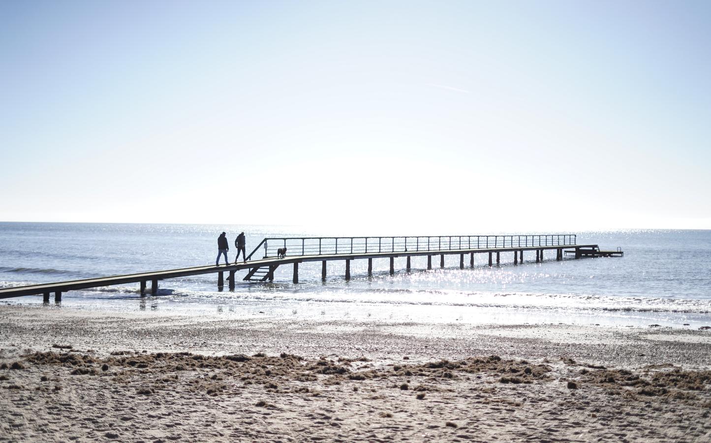 Par som går på stranden på vesterhavet ved en badebro